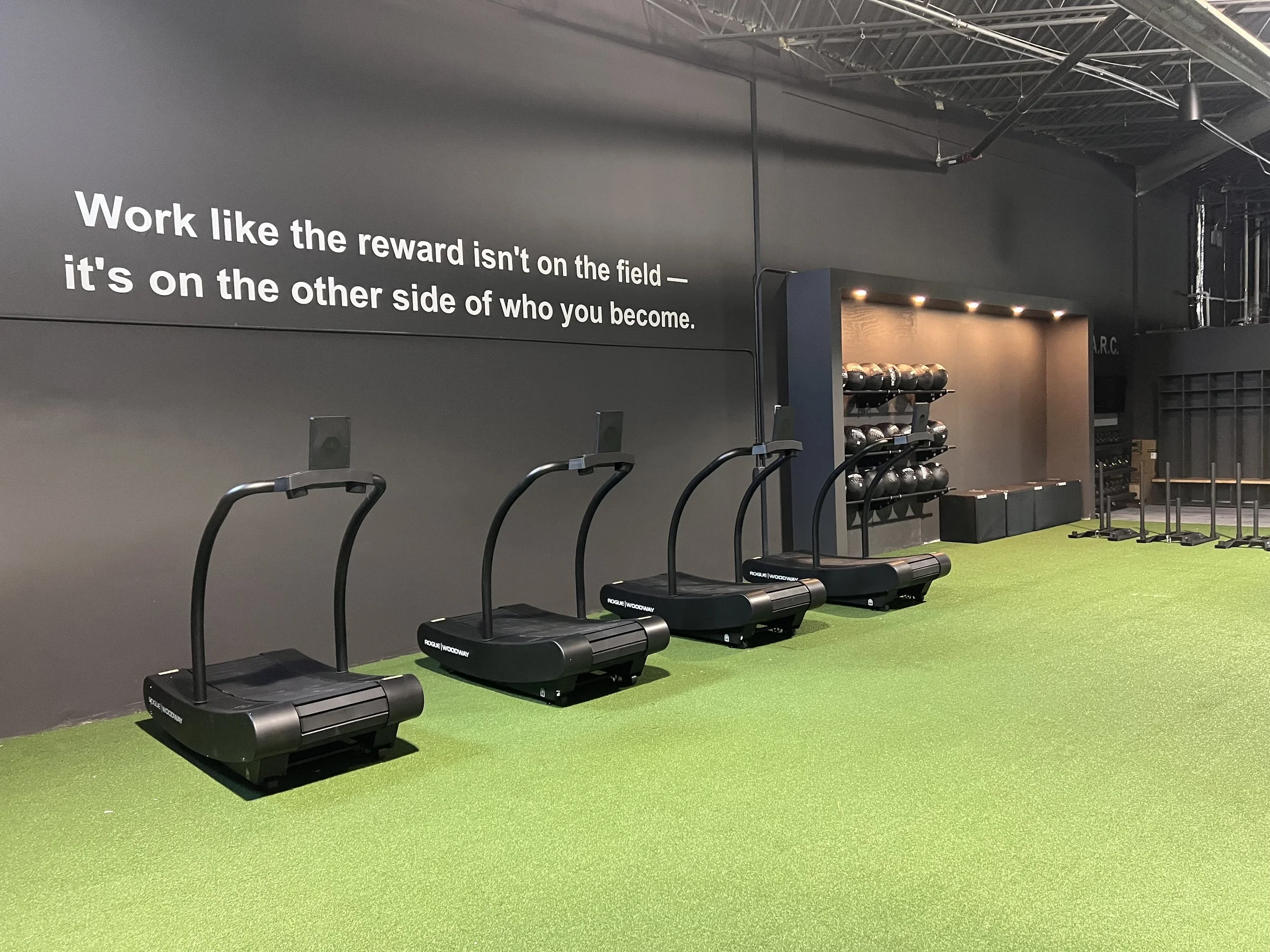 Inside a gym with four treadmills on green rubber flooring, a wall with motivational quote, shelves with kettlebells, and workout equipment.