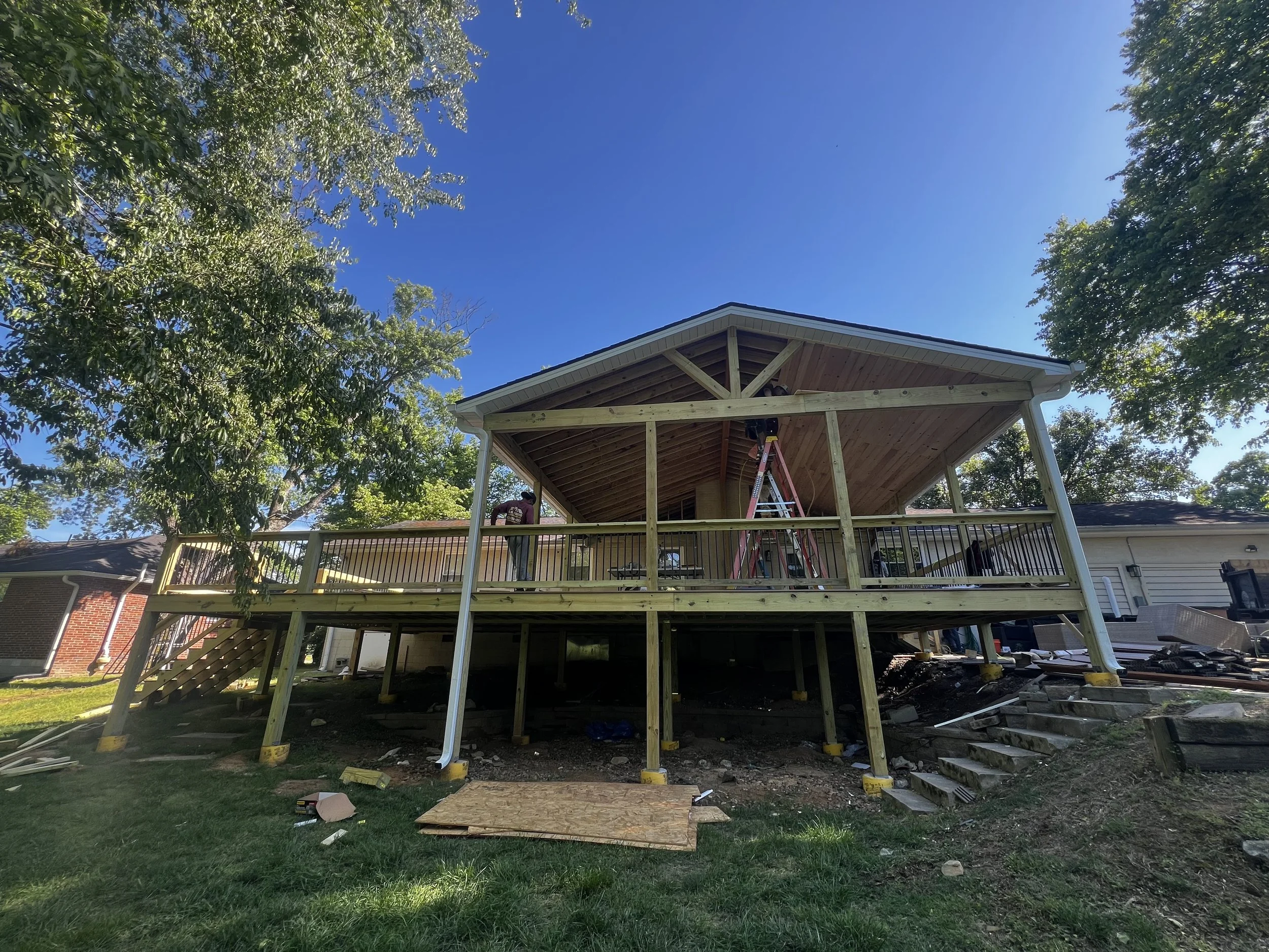 A partly constructed wooden deck attached to a house, with workers building it. The deck has a roof, and safety railings are in place. Construction tools and materials are scattered below the deck. The surrounding area includes trees, grass, and neighboring houses, and the sky is clear and blue.