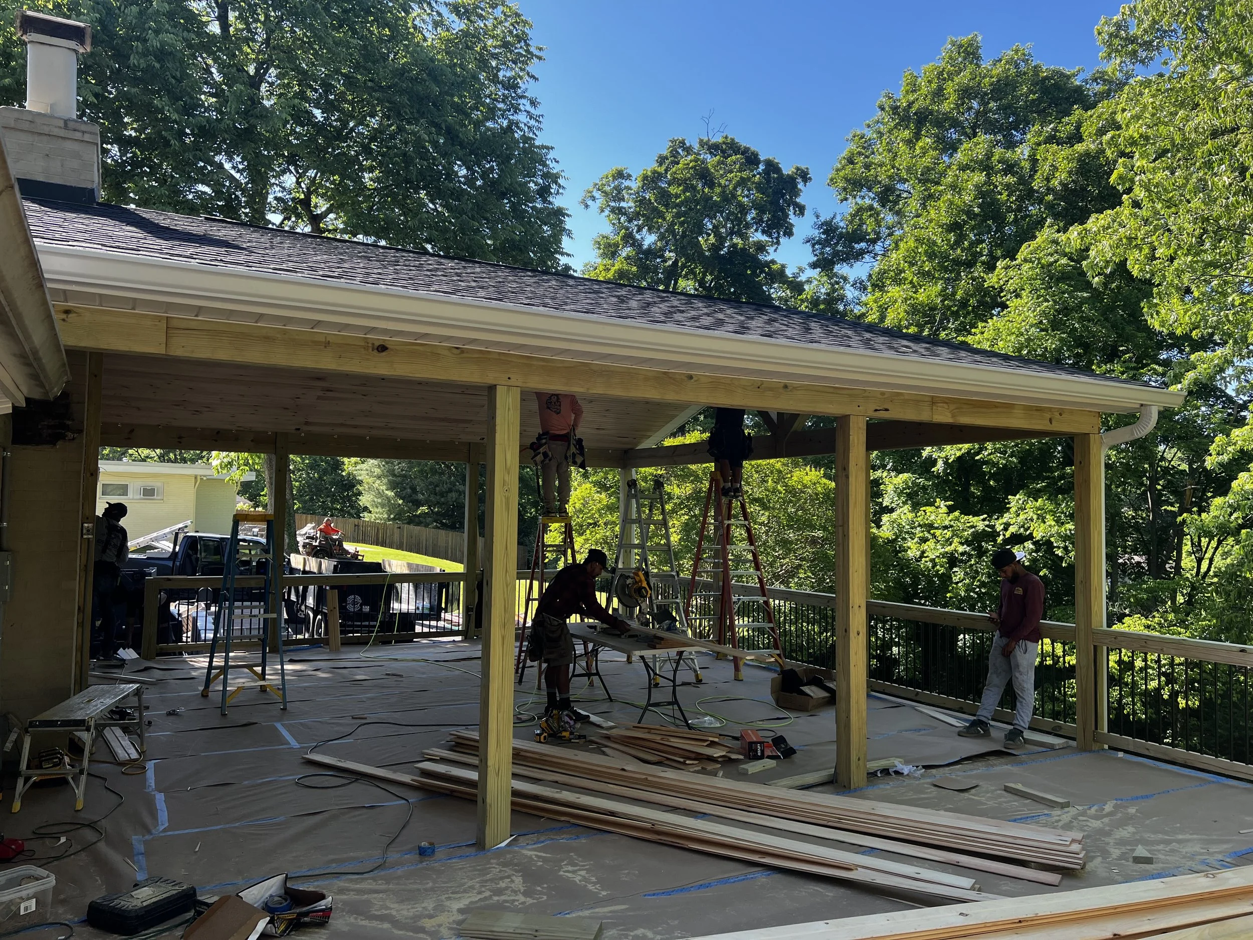 Construction workers building a wooden deck in a backyard, with tools, wood planks, and ladders visible on the floor. Green trees and a clear blue sky are in the background.