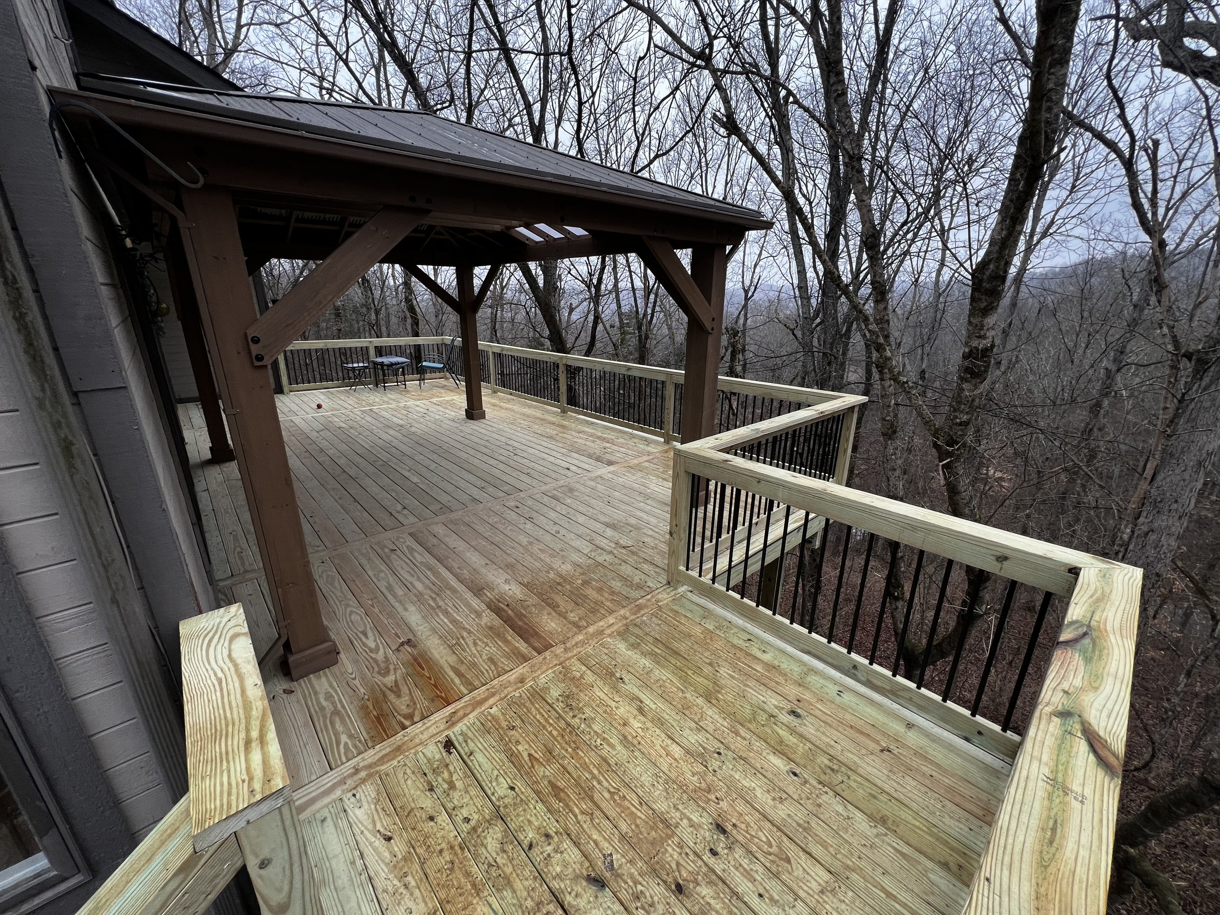 A wooden deck with a railing in a wooded area during daytime, with trees without leaves in the background.