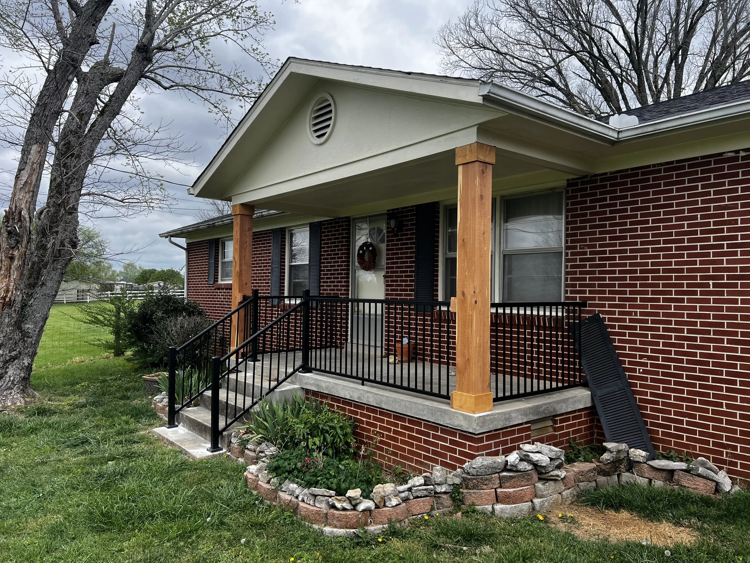 The image shows a house with a brick exterior and a front porch with black railings. The porch has wooden posts supporting the roof, and there are steps leading up to the porch. A wreath decorates the front door. The yard is grassy with a small stone-edged flower bed in front of the porch, containing green plants and flowers. Two large leafless trees are on the left and right sides of the house, and the sky is overcast with gray clouds.