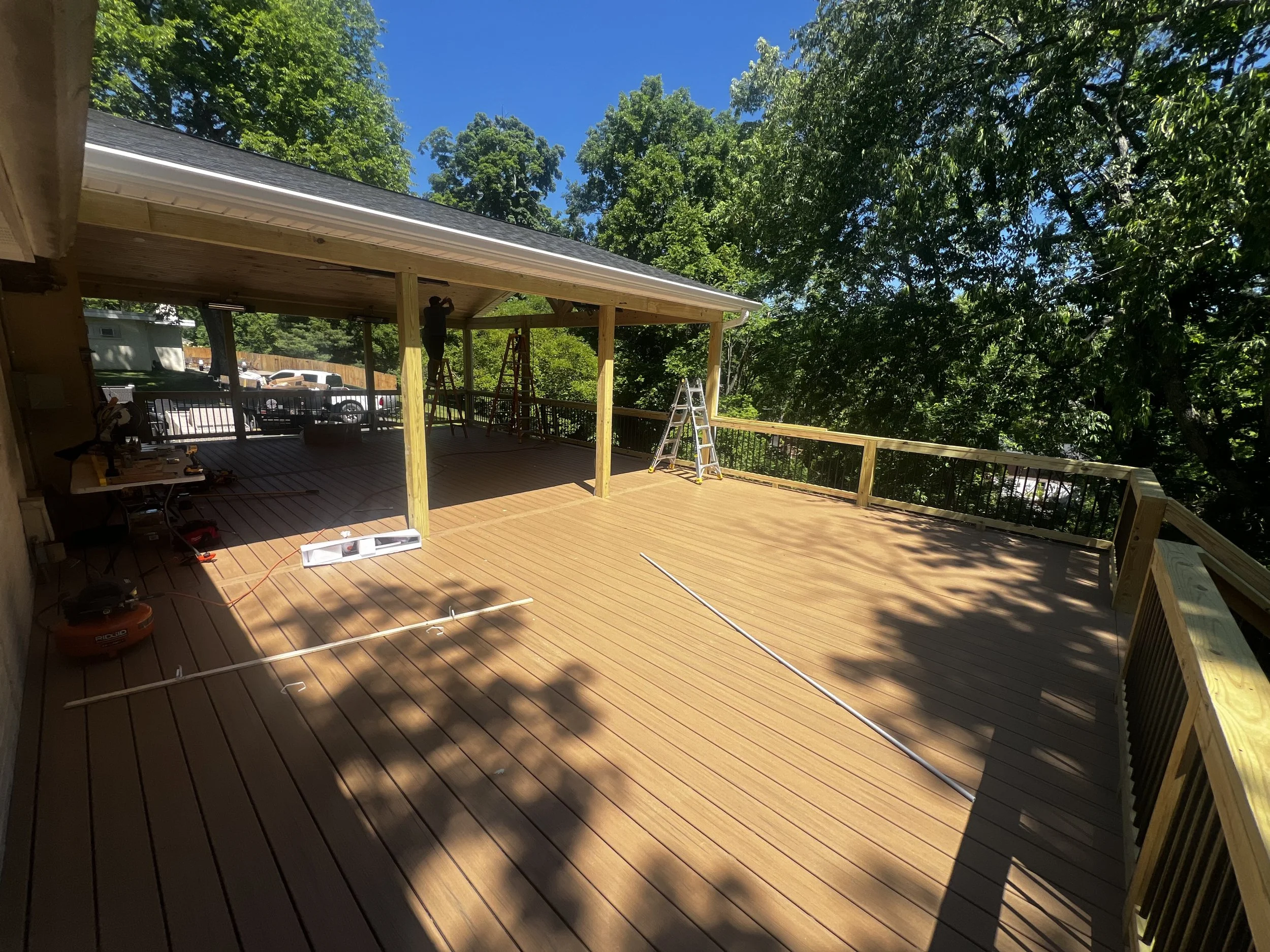 People installing a wooden deck on a house's porch, with ladders and tools, surrounded by trees under a blue sky.