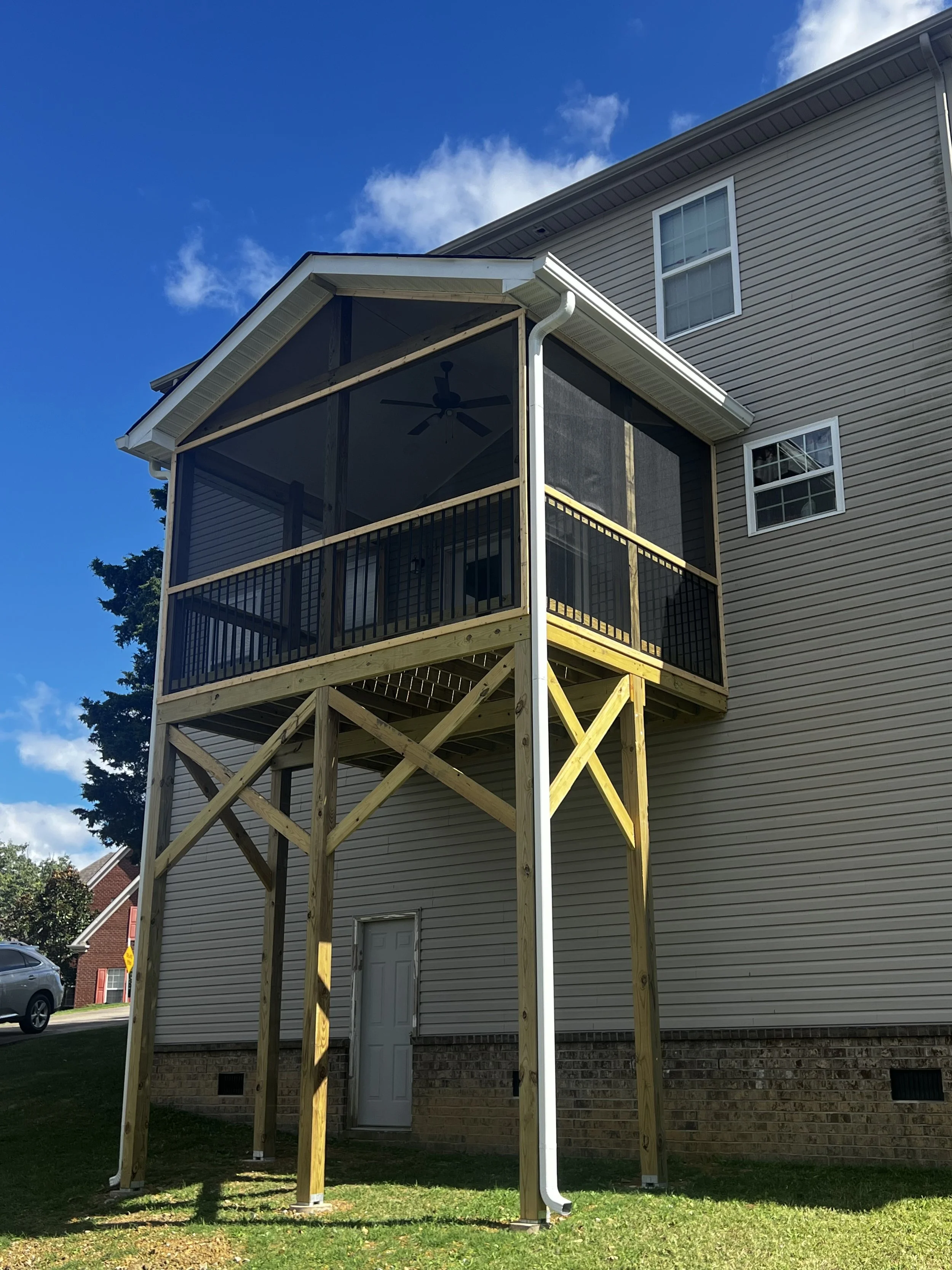 A multi-story house with a screened-in porch on the second floor. The porch has black railing and wooden support beams. The house has beige siding with white trim, and an outdoor ceiling fan is visible inside the porch. A white downspout runs along the corner of the house, and a white door is on the first floor. The sky is blue with some clouds, and there is a car and neighboring houses in the background.