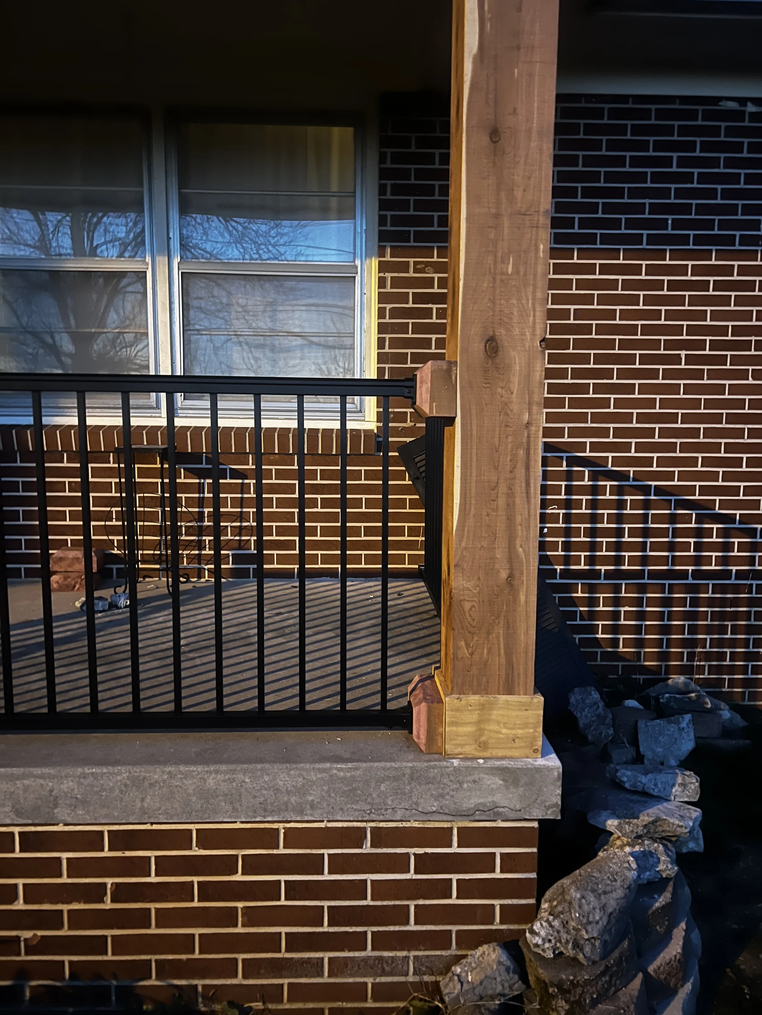 Close-up of a brick building with a window, a metal railing, and a wooden post, with rocks on the ground in front.