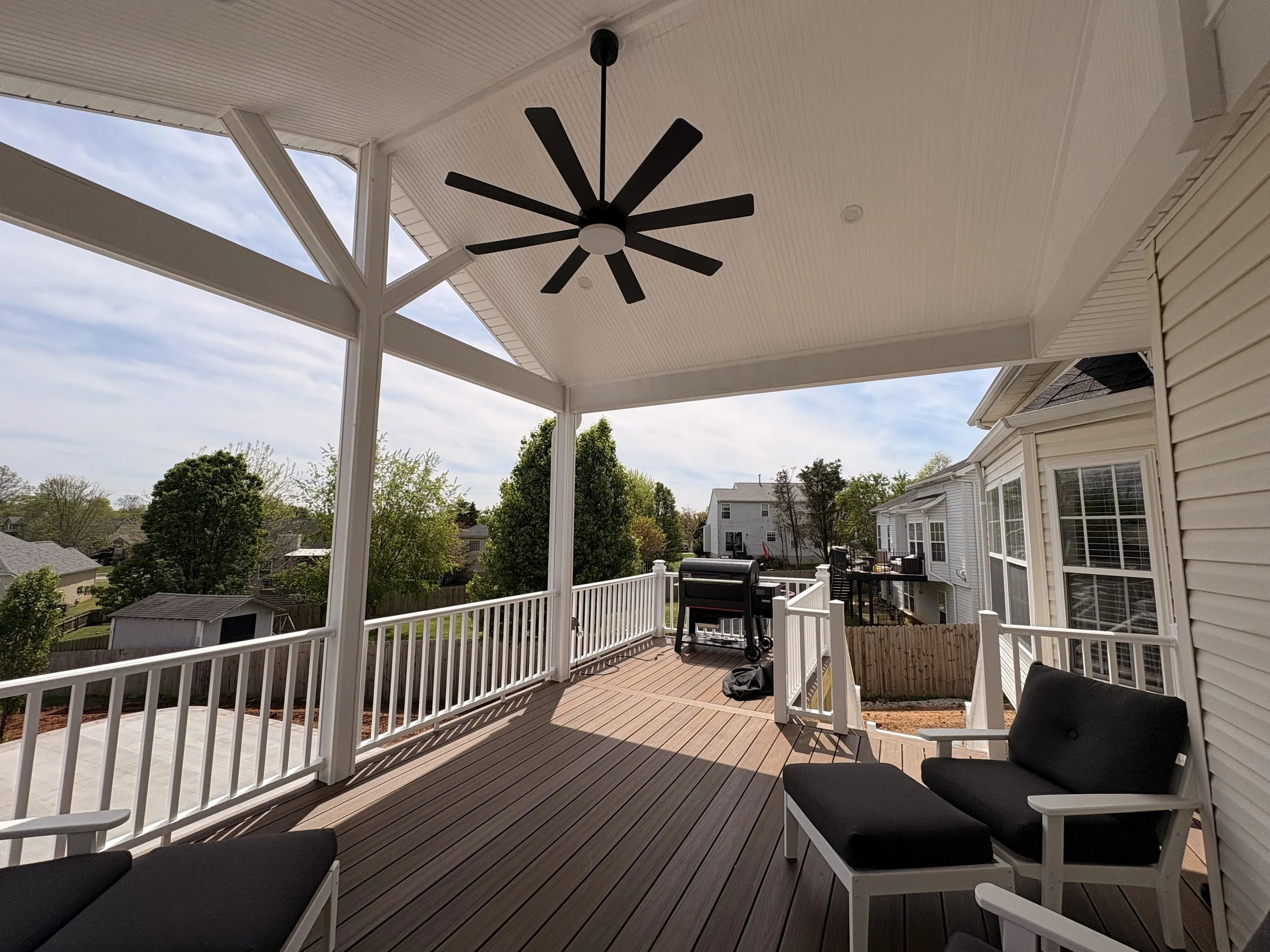 A covered outdoor porch with a ceiling fan, seating area with cushions, a barbecue grill, and view of surrounding houses and trees under a partly cloudy sky.