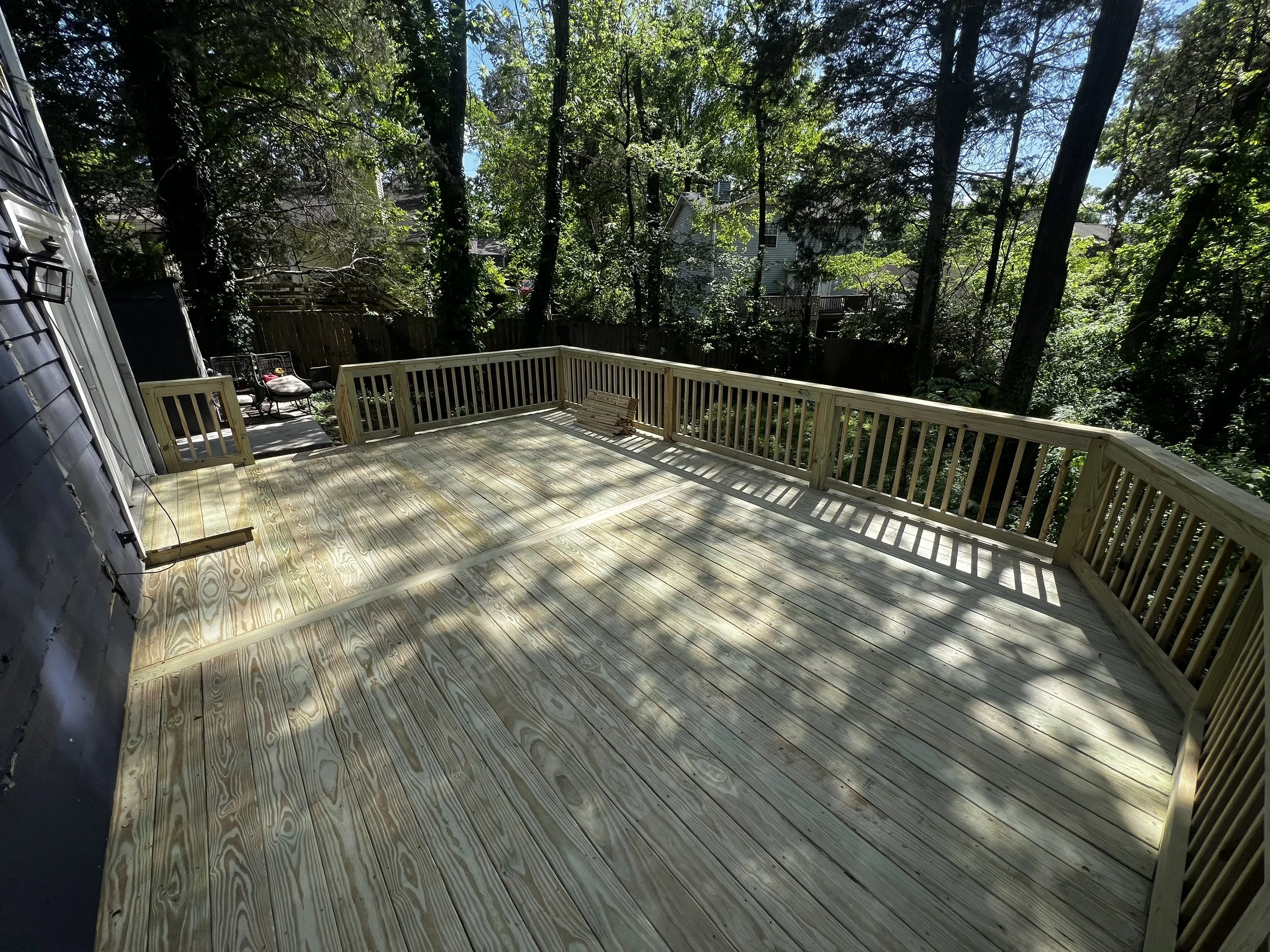 A newly built wooden deck with railing, surrounded by tall trees and neighboring houses visible in the background.