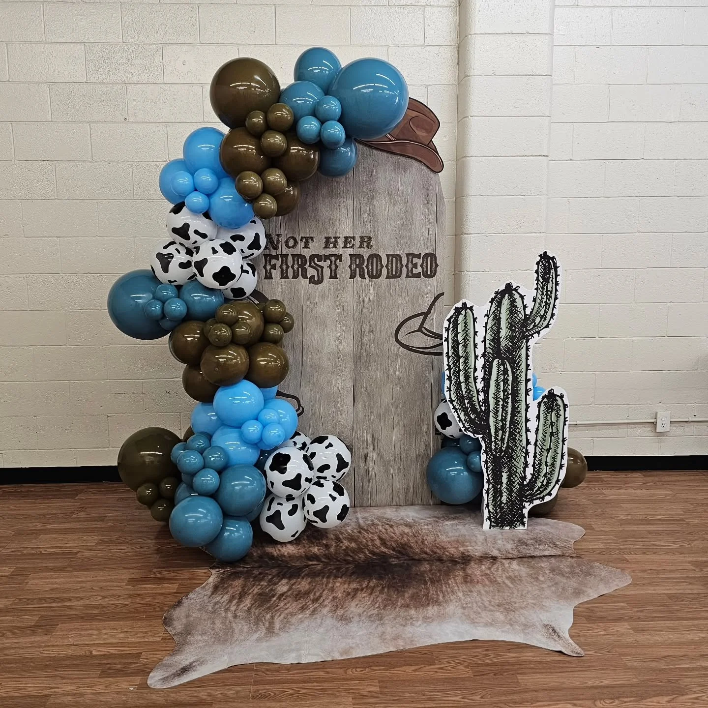 🤠✨ Not Her First Rodeo! This western backdrop is giving all the rustic cowgirl vibes &mdash; cow print balloons, earthy browns, pops of blue, a cactus prop, and the perfect cowhide rug to tie it all together. The ultimate photo spot for any rodeo-th