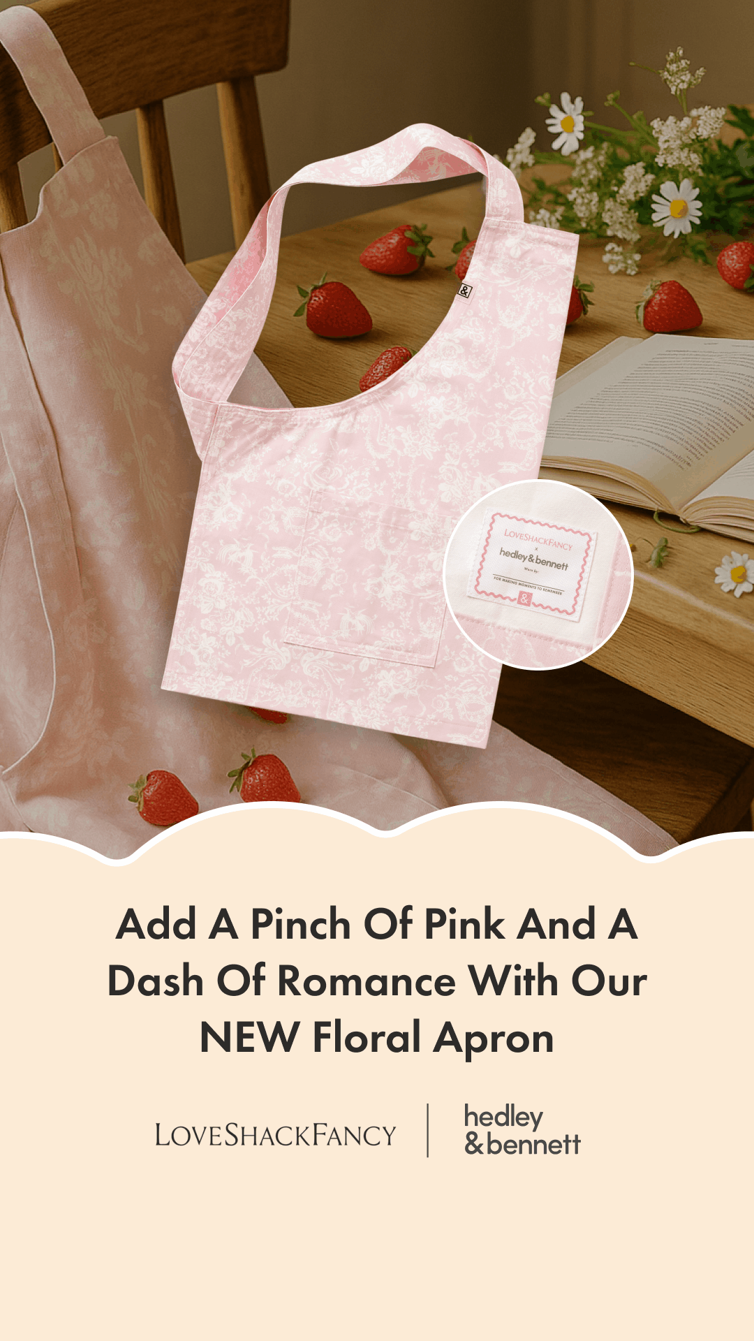 A pink floral apron with a logo tag, surrounded by strawberries and daisies on a wooden table, with an open book nearby.
