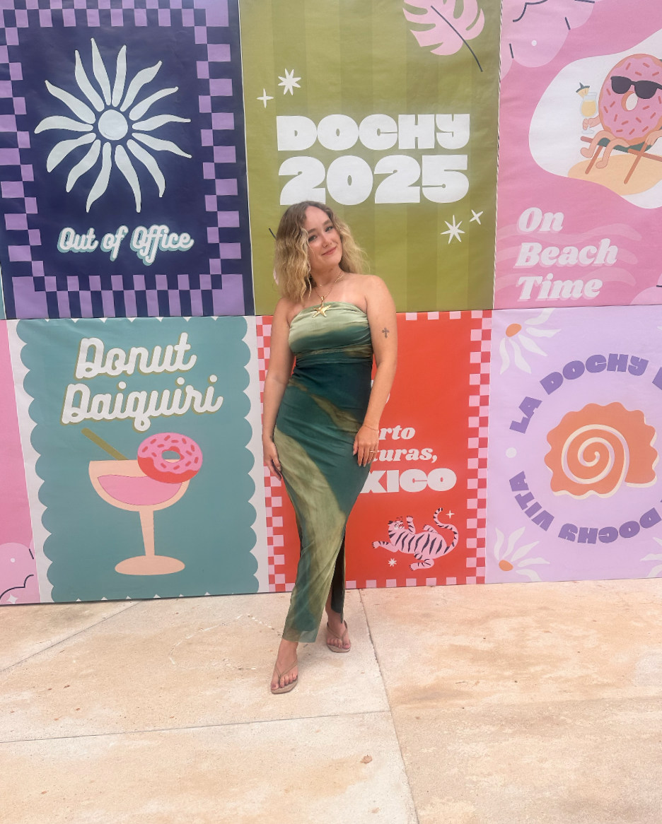 A woman in a strapless green and beige dress standing in front of a colorful backdrop with various summer and beach-themed designs and text.