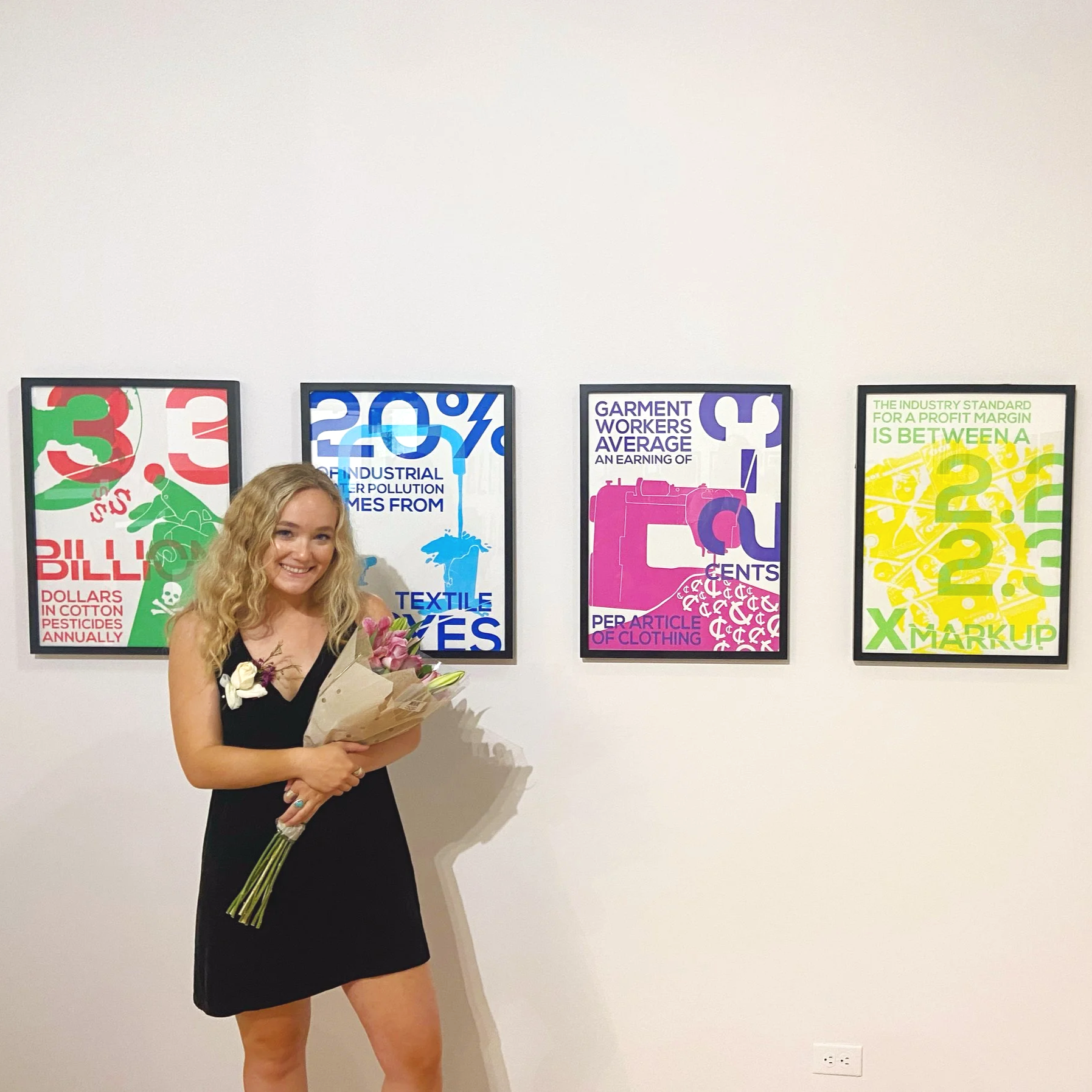 Woman with curly blonde hair in a black dress holding a bouquet of flowers, standing in front of colorful posters on a white wall.