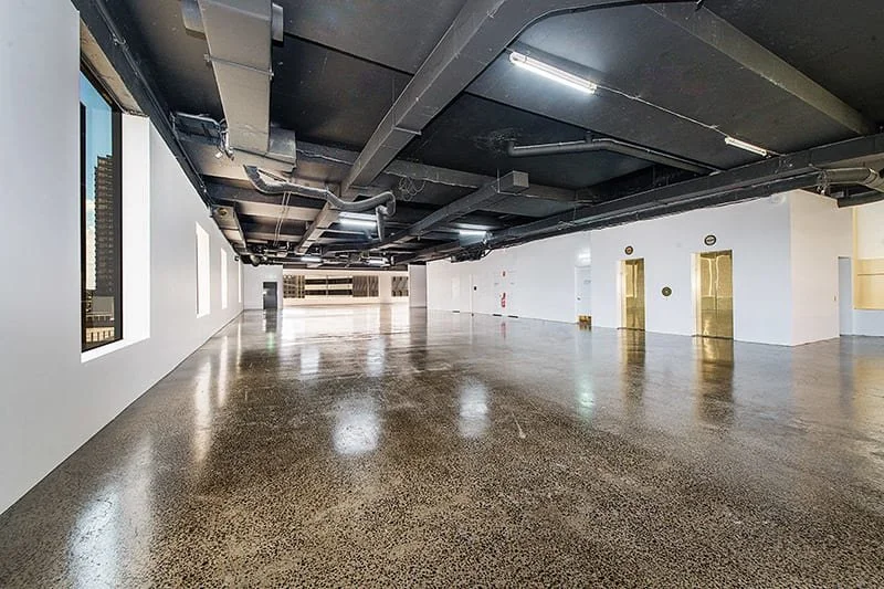 Empty commercial space or office with polished concrete floors, white walls, large windows, and exposed black ceiling with ductwork and lighting.