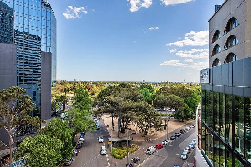 Cityscape view with modern glass buildings, parking lot, and green trees under a blue sky with scattered clouds.
