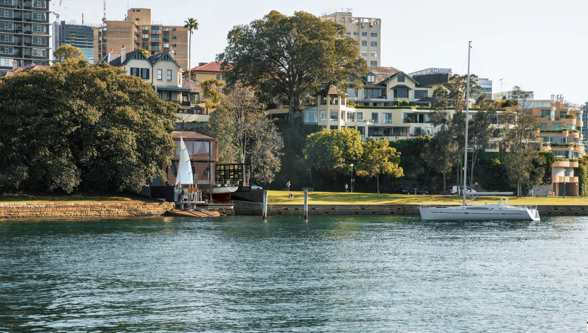 A waterfront scene with a sailboat on the water, a grassy area with trees and people, and various residential buildings on a hillside in the background.