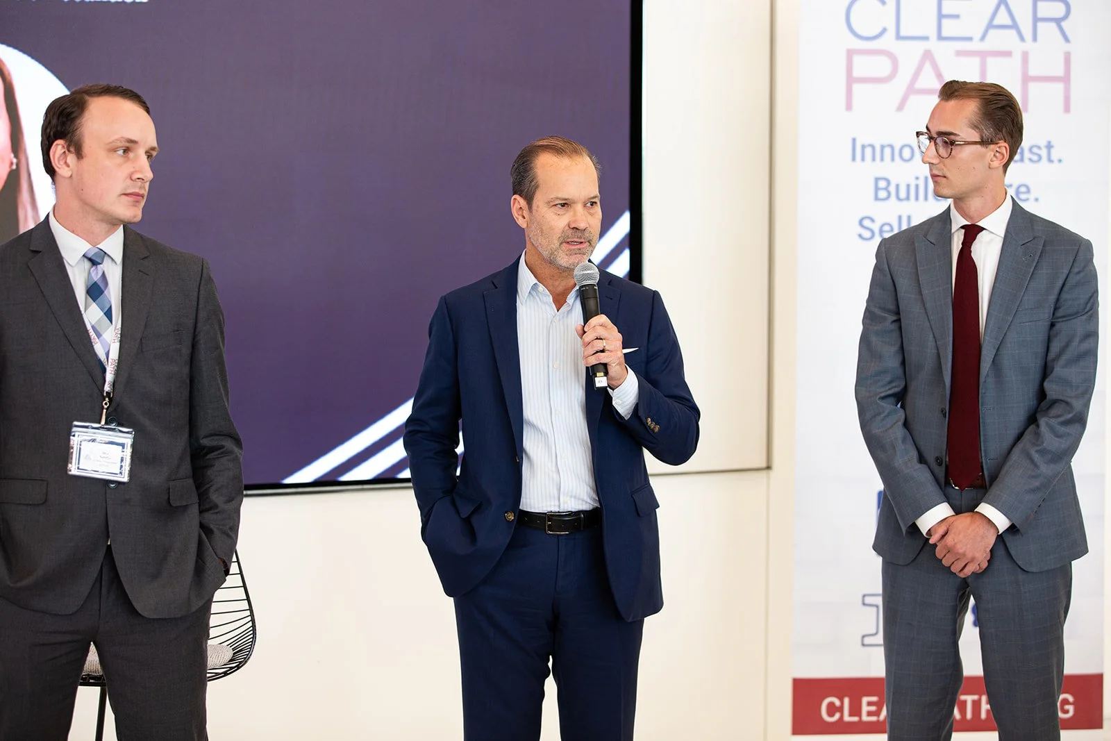 Three executives participate in a discussion at a corporate event, with the featured speaker addressing attendees while standing between fellow presenters. Captured during a professional conference session, the image highlights executive leadership, 