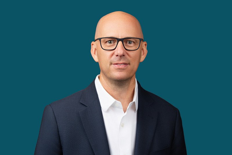 A professional headshot featuring a business executive dressed in a navy suit and white shirt against a clean, modern teal background. The portrait captures a confident and approachable presence, highlighting the polished style and professional image