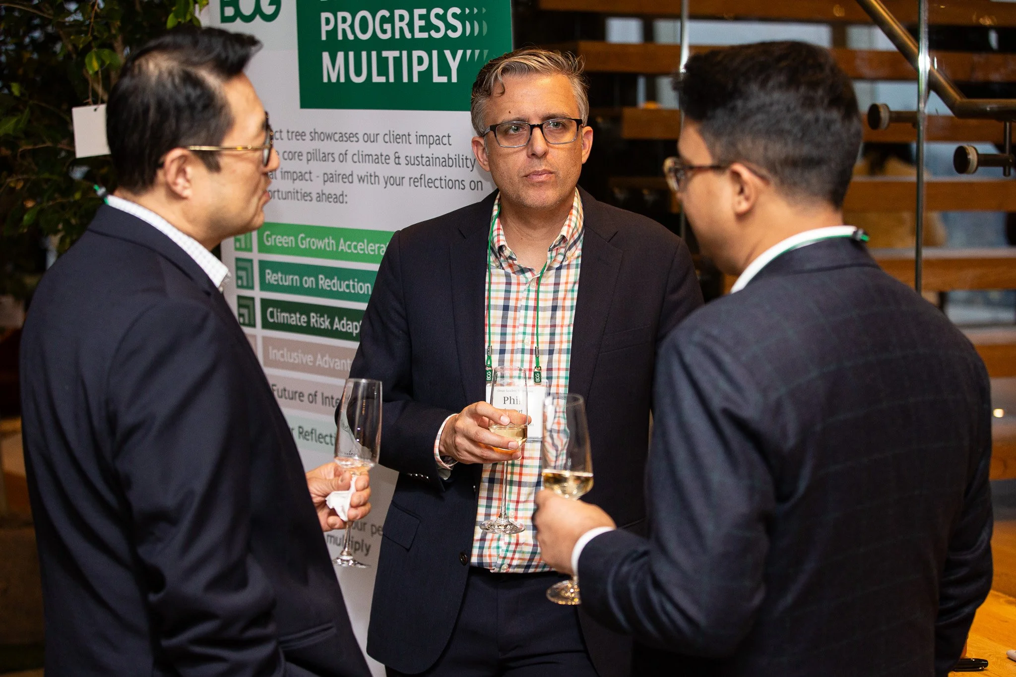Three men in business attire having a conversation at a networking event, with a poster about climate impact and sustainability in the background.