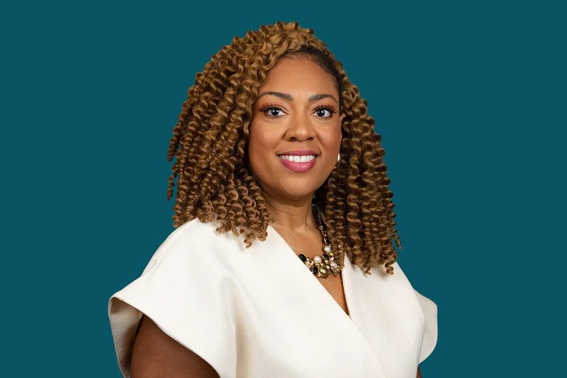 A professional headshot featuring a business executive in a white outfit against a clean teal background. The portrait highlights a confident, polished presence ideal for corporate branding. 