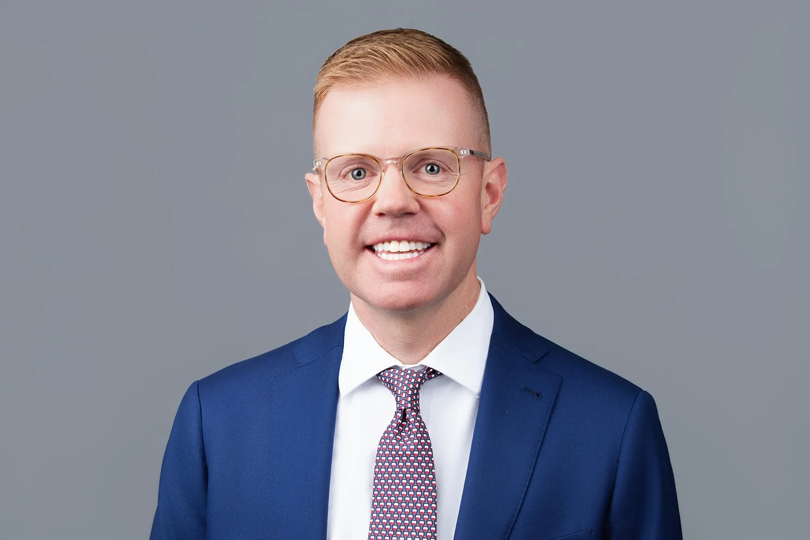 Portrait of a smiling man with glasses, wearing a blue suit, white shirt, and patterned tie, against a gray background.