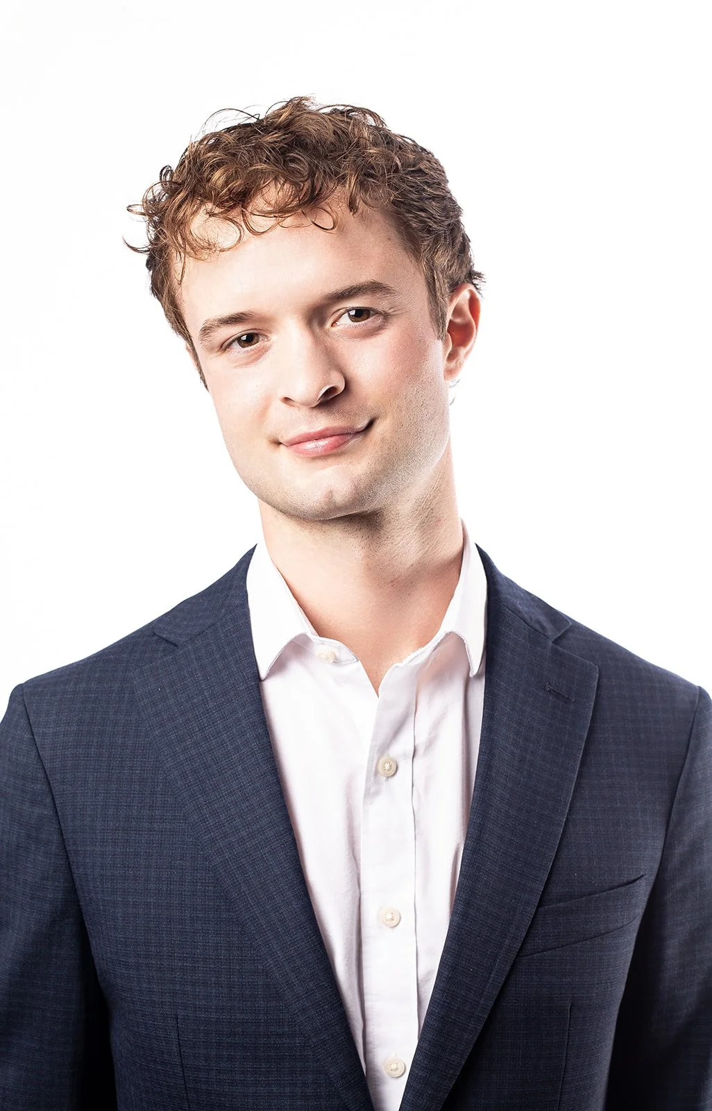 Portrait of a young man with curly brown hair wearing a dark suit and white shirt, smiling gently at the camera.
