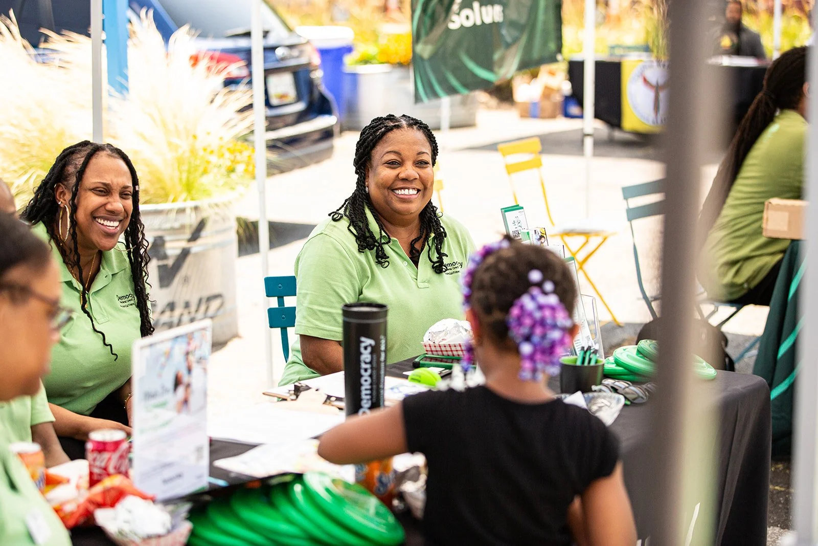 Women in green shirts sitting at a table smiling and talking with a young girl with purple braids at an outdoor event.