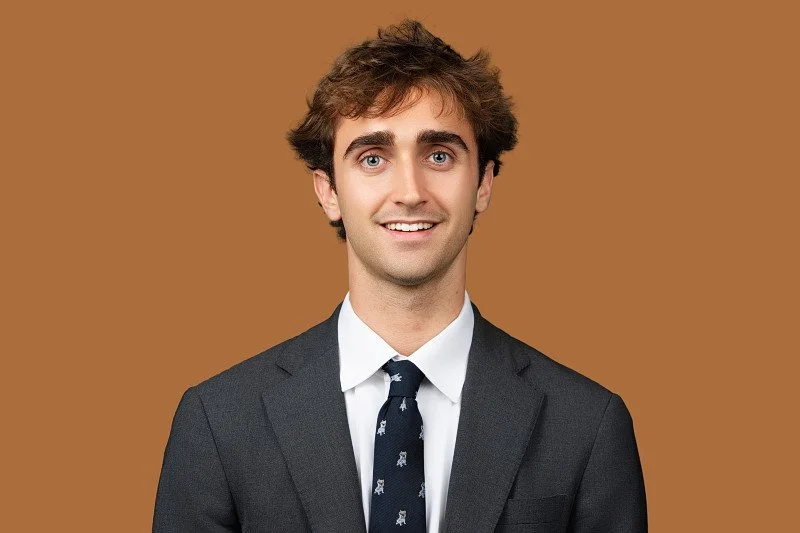 A professional headshot of a young man wearing a charcoal gray suit, white dress shirt, and navy patterned tie, smiling at the camera against a warm brown background.