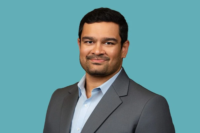 A professional headshot of a smiling man with short dark hair wearing a gray suit jacket and light blue dress shirt, photographed against a soft teal background.