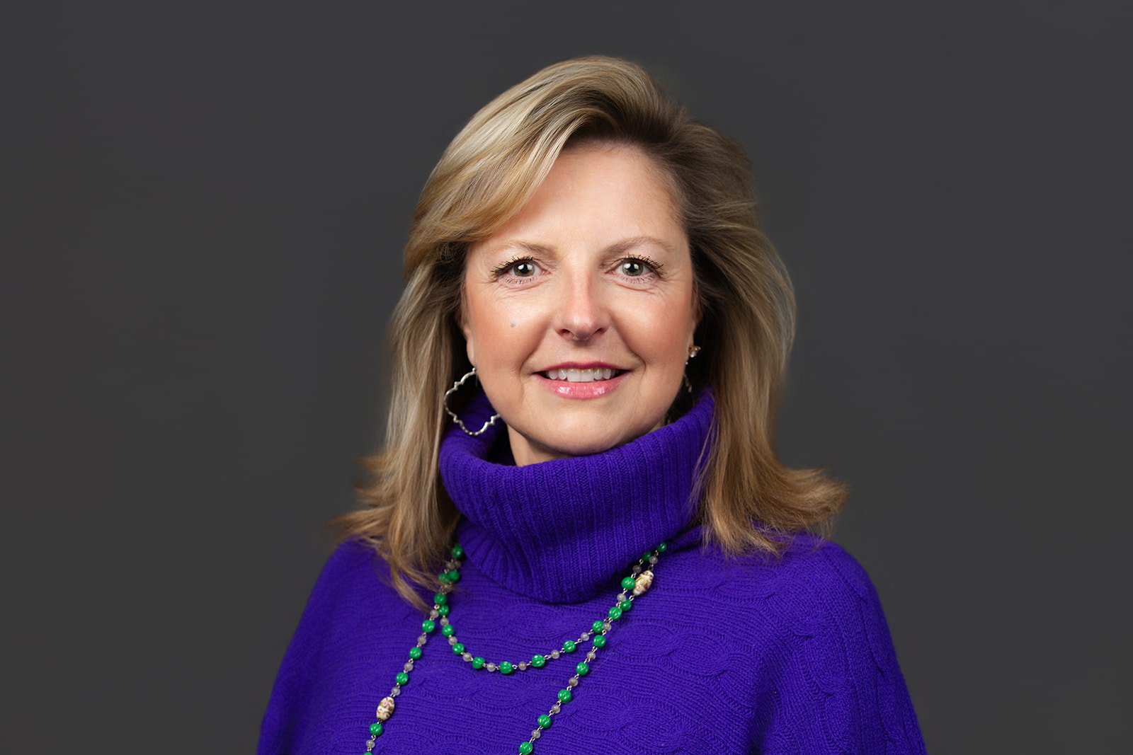 A woman with shoulder-length blonde hair, smiling, wearing a purple turtleneck sweater, silver hoop earrings, and a green beaded necklace against a gray background.