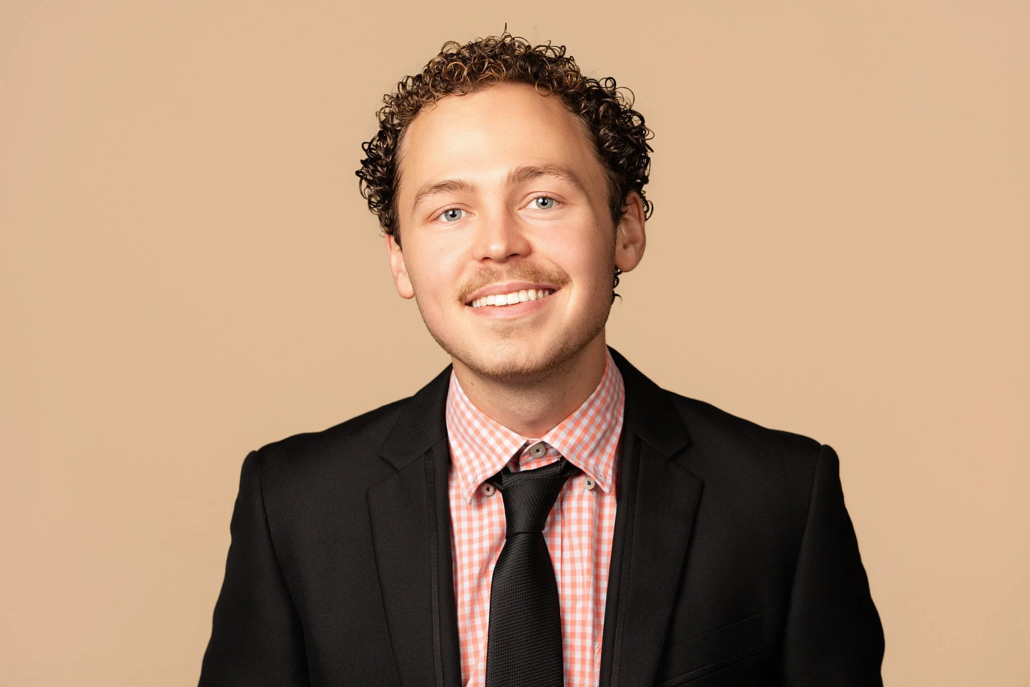 Portrait of a young man with curly hair, wearing a black blazer, a pink checkered shirt, and a black tie, smiling against a beige background.