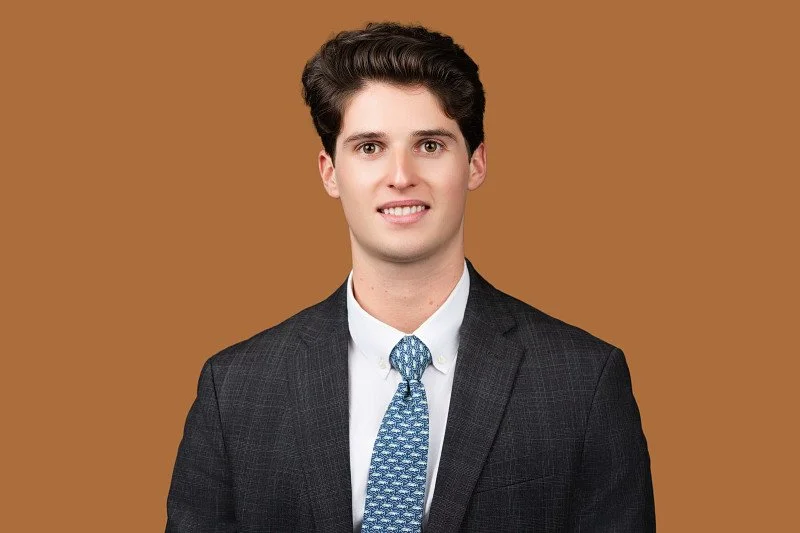 A professional headshot featuring a man wearing a dark suit, white shirt, and patterned blue tie against a warm brown background.