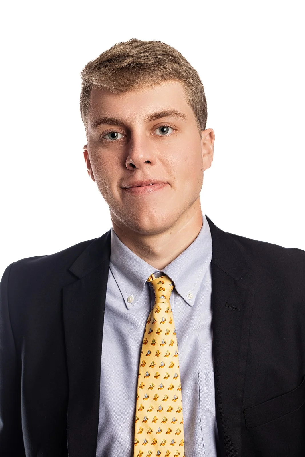 Portrait of a young man in a dark suit, light blue shirt, and yellow tie with small pattern, against a white background.