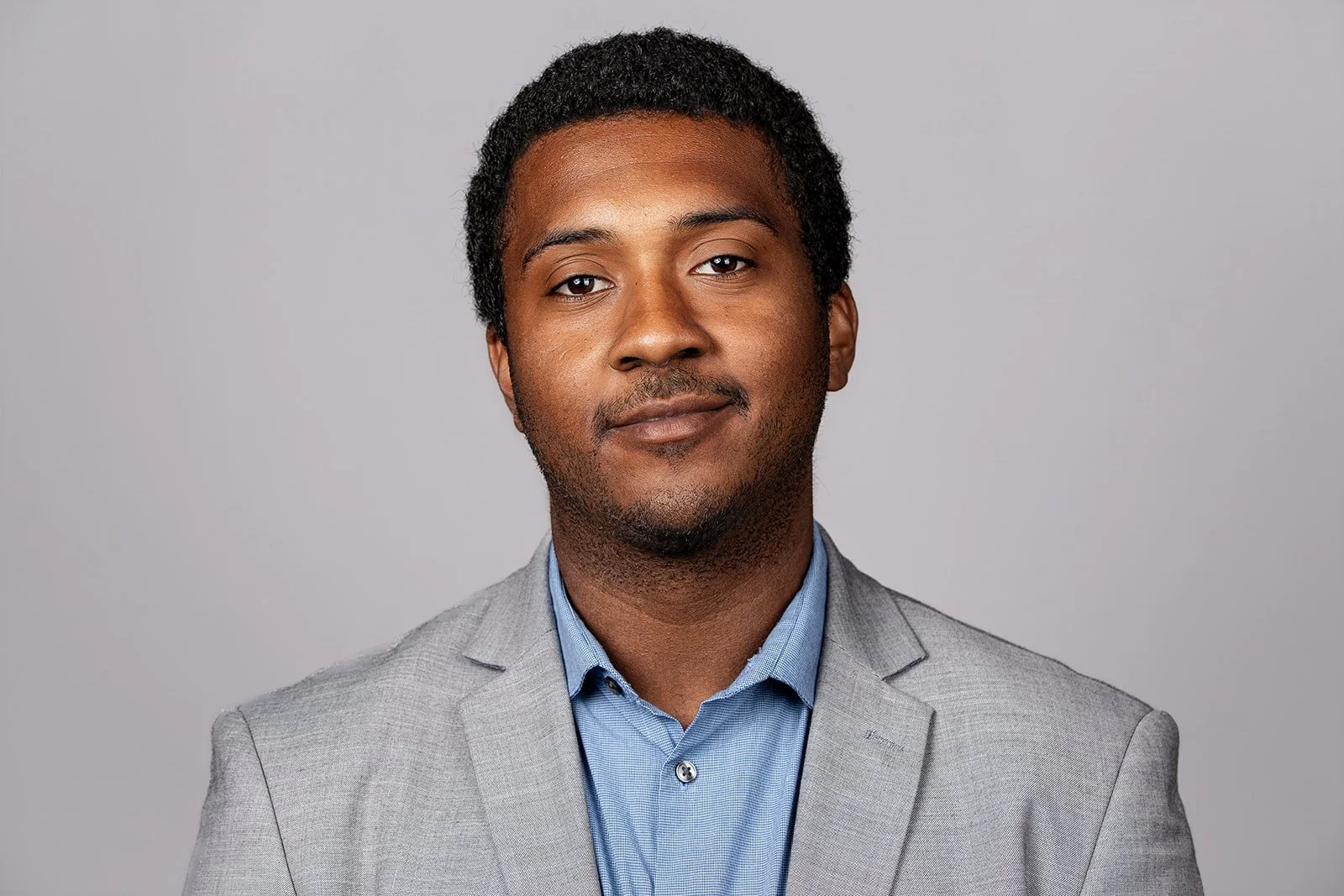 Portrait of a young man wearing a light gray blazer and a blue shirt, posing against a plain light gray background.