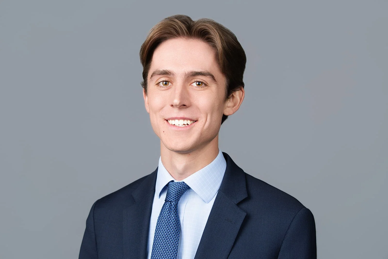 Portrait of a young man in a dark suit and blue tie, smiling against a plain gray background.