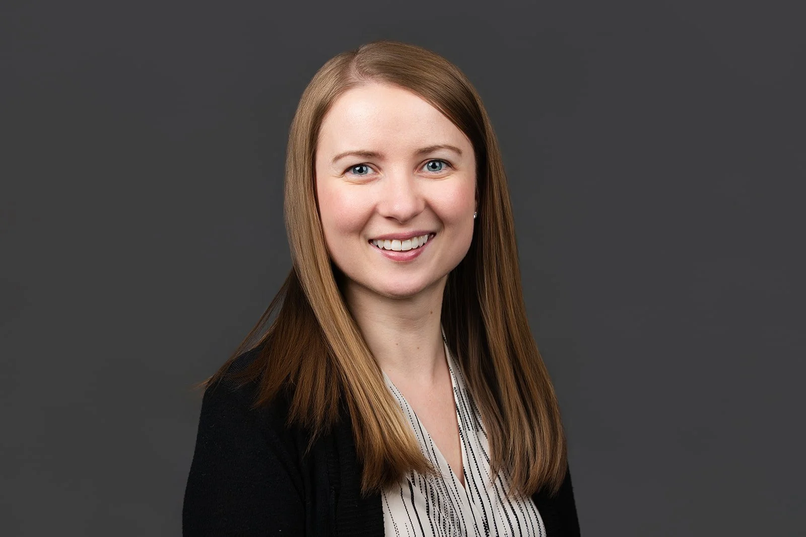 Headshot of a woman with long straight hair, blue eyes, smiling, wearing a black blazer and a striped blouse, against a gray background.