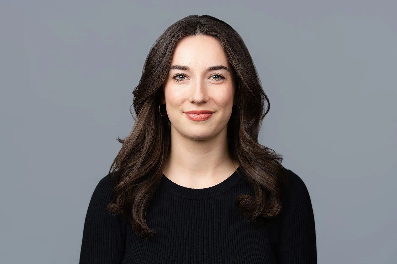 Headshot of a woman with long wavy brown hair, wearing a black top and hoop earrings, smiling lightly, against a plain gray background.