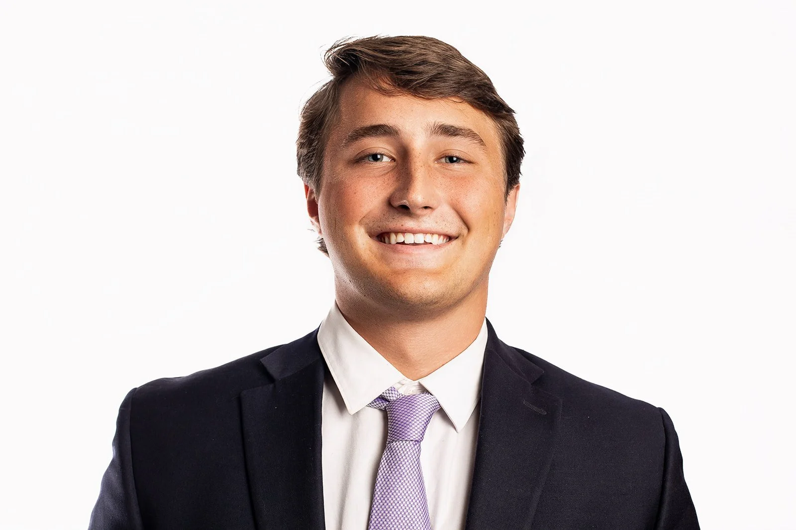 A young man with light brown hair, wearing a black suit jacket, white shirt, and lavender tie, smiling against a white background.