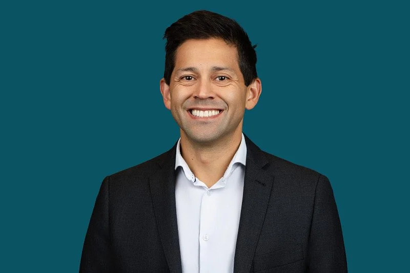 A professional headshot featuring a man wearing a dark blazer and white shirt against a deep teal background. The portrait showcases a confident smile.