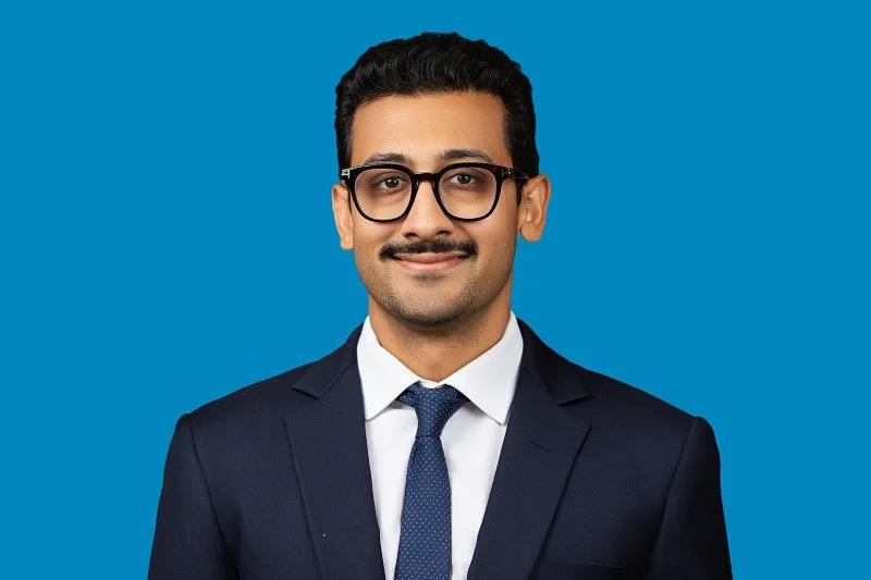 A professional headshot featuring a man wearing a navy suit, white shirt, and tie against a vibrant blue background. The portrait showcases a polished appearance and confident expression.