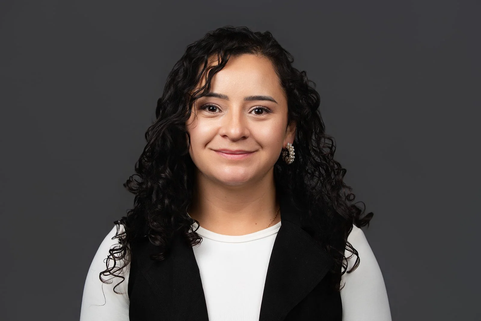 Portrait of a woman with dark curly hair, wearing earrings, a white top, and a black vest, smiling against a dark gray background.