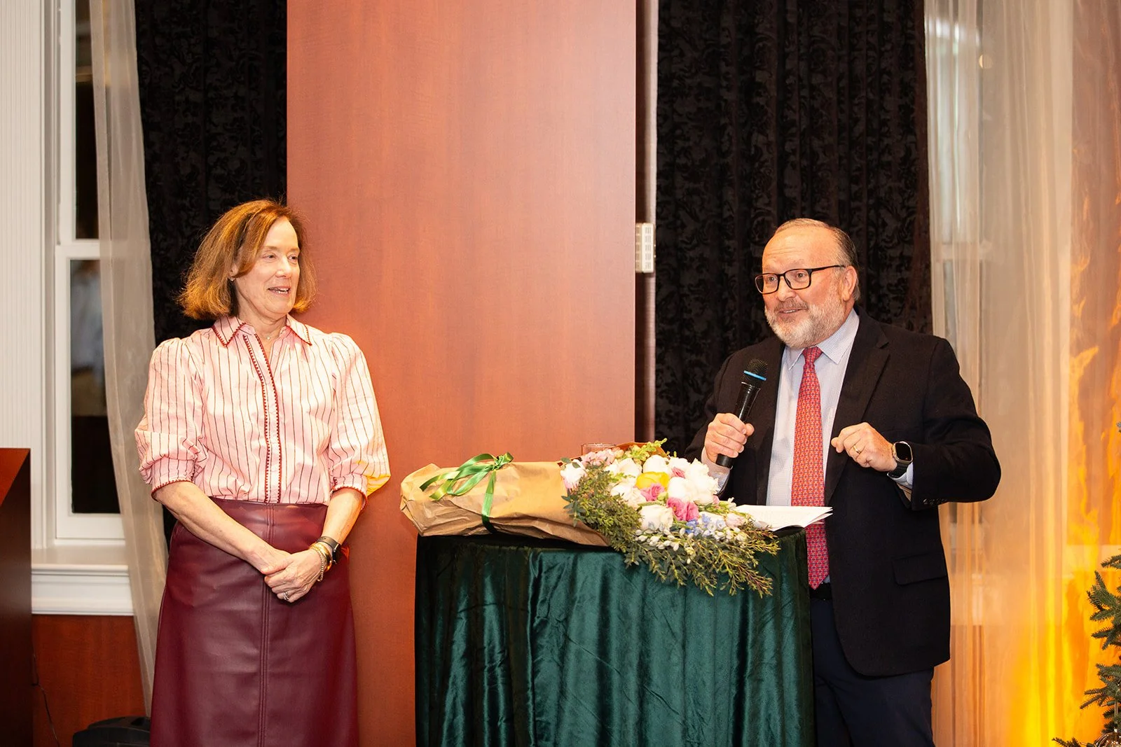 A woman and a man at a formal event. The woman is standing and smiling, wearing a striped blouse and a maroon skirt. The man, wearing glasses, a suit, and a red tie, is speaking into a microphone and standing behind a table with a bouquet of flowers.