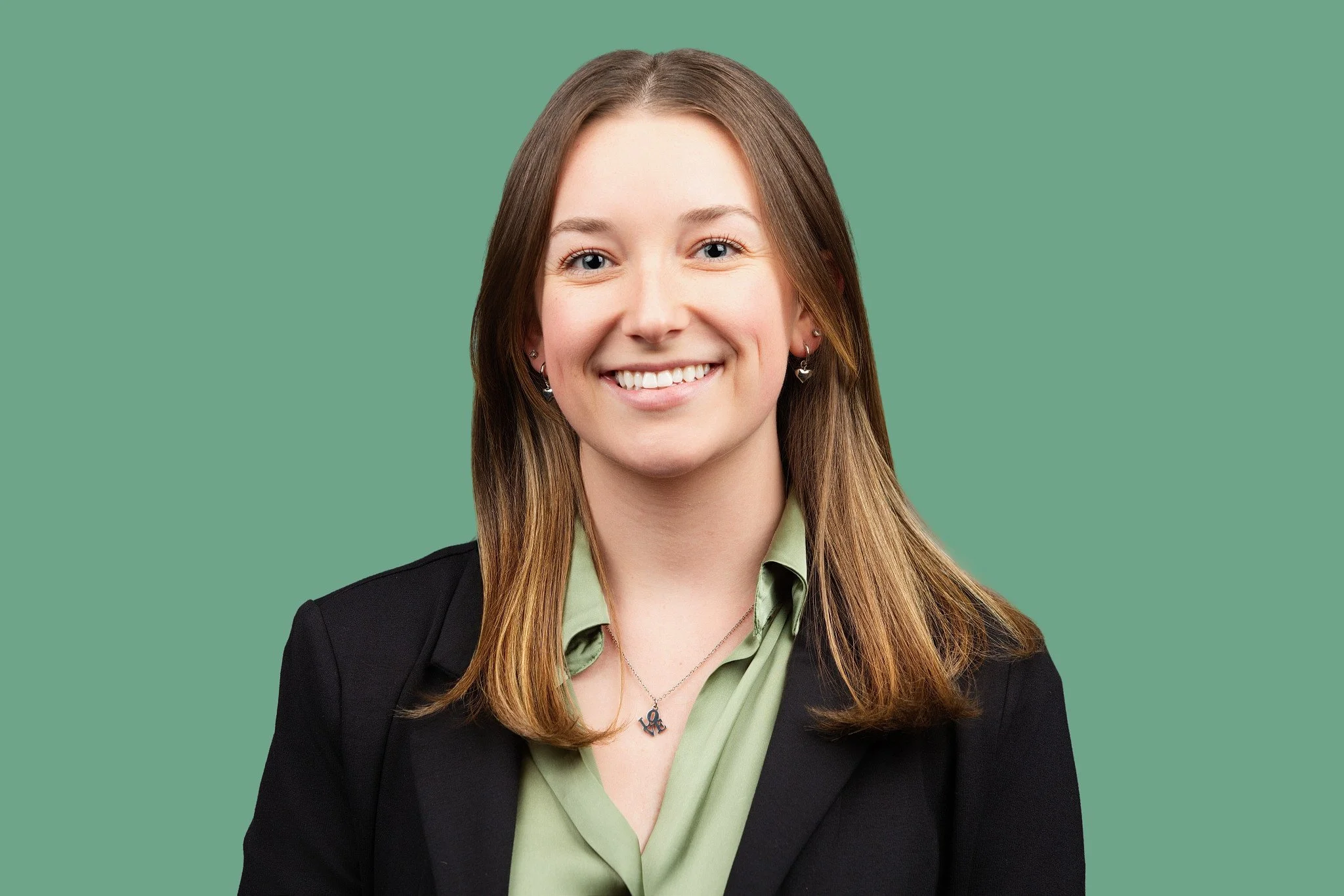 A young woman with long straight brown hair with subtle highlights is wearing a black blazer over a soft sage green blouse, along with small drop earrings and a delicate pendant necklace, smiling broadly, posed against a muted green background. 