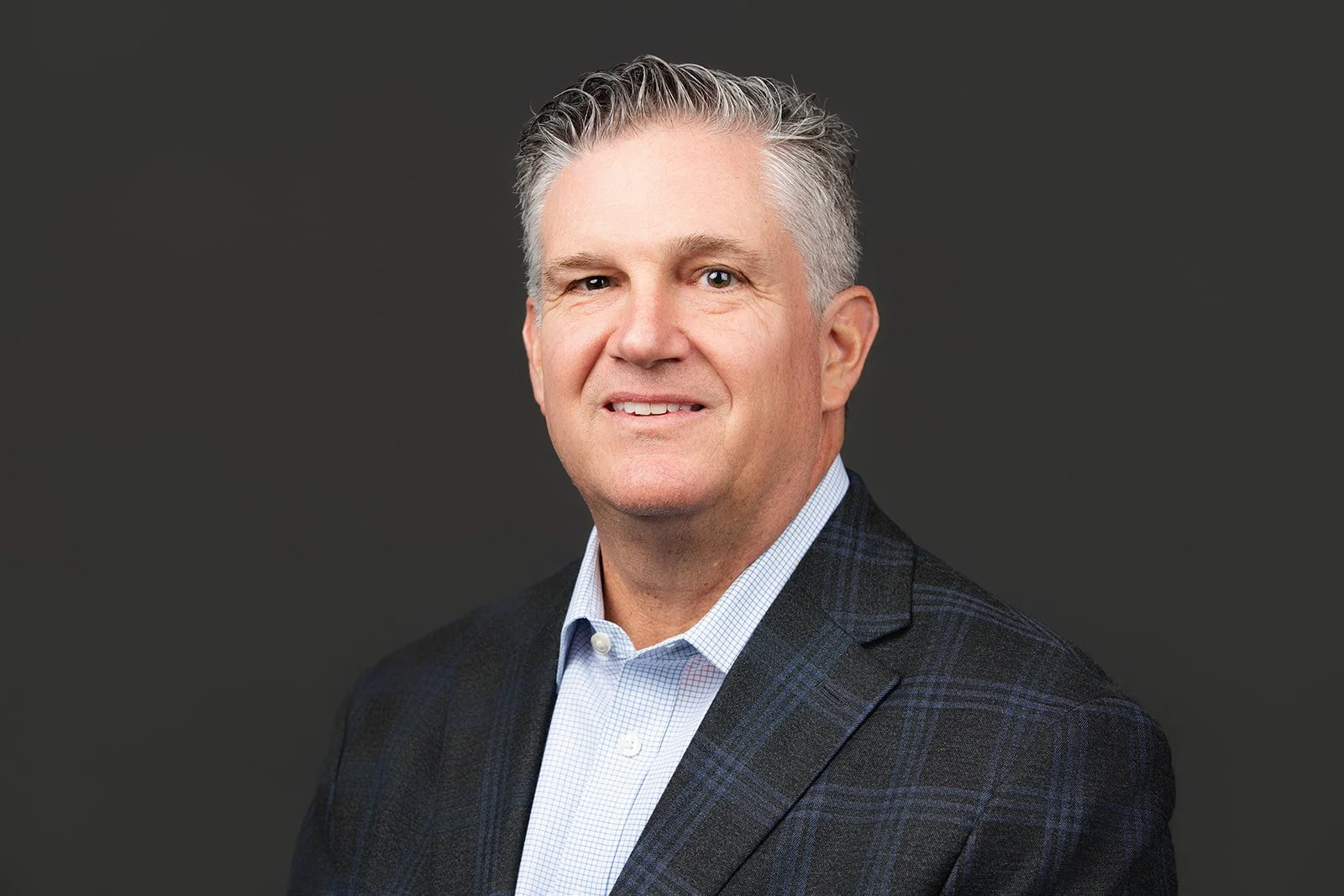 A middle-aged man with gray hair, wearing a checkered blazer and light blue dress shirt, smiling slightly against a dark gray background.