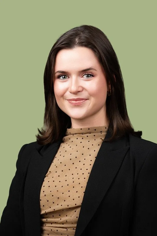 A professional headshot of a smiling woman with shoulder-length dark brown hair wearing a black blazer over a tan polka dot blouse, photographed against a soft sage green background.