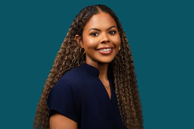 A professional headshot of a smiling woman with long curly hair wearing a navy blouse, photographed against a deep teal background.