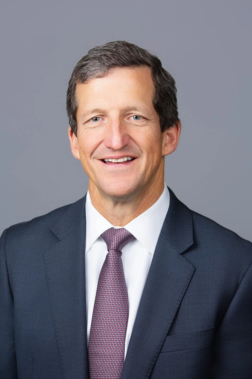 A professional headshot of a middle-aged man in a dark suit, white shirt, and a patterned tie, set against a plain gray background.