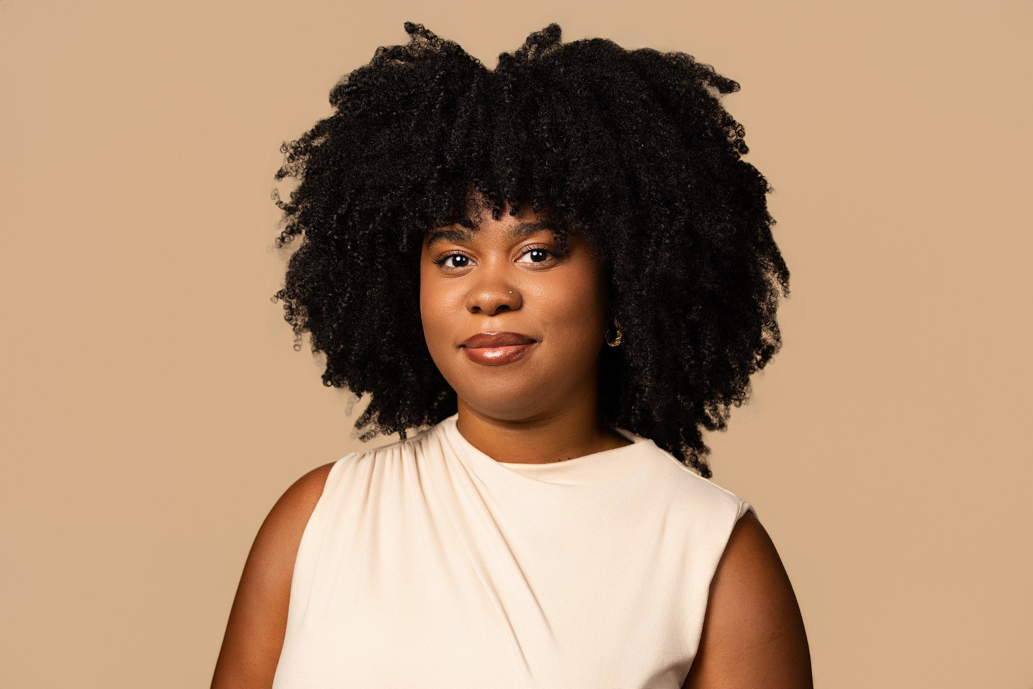 Portrait of a Black woman with natural curly hair wearing a sleeveless beige top standing against a beige background.