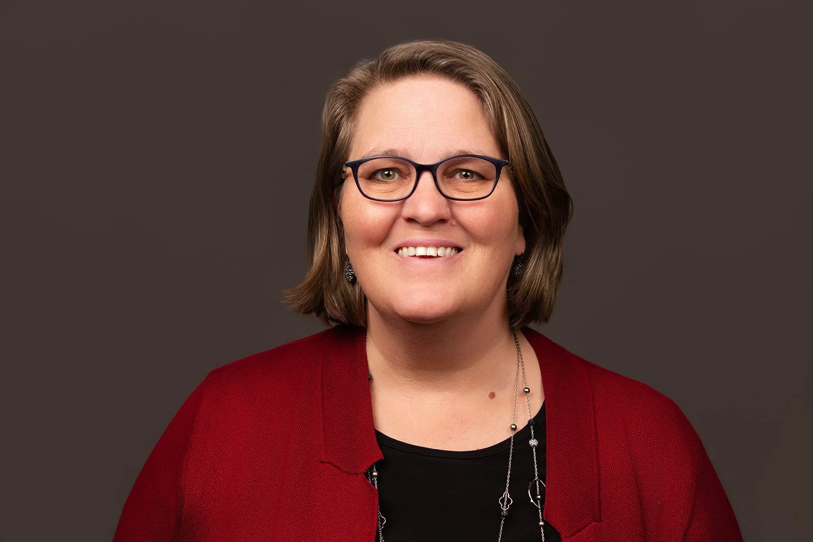 A portrait of a woman with shoulder-length brown hair, wearing glasses, a black top, a red blazer, and a silver necklace, smiling against a dark gray background.