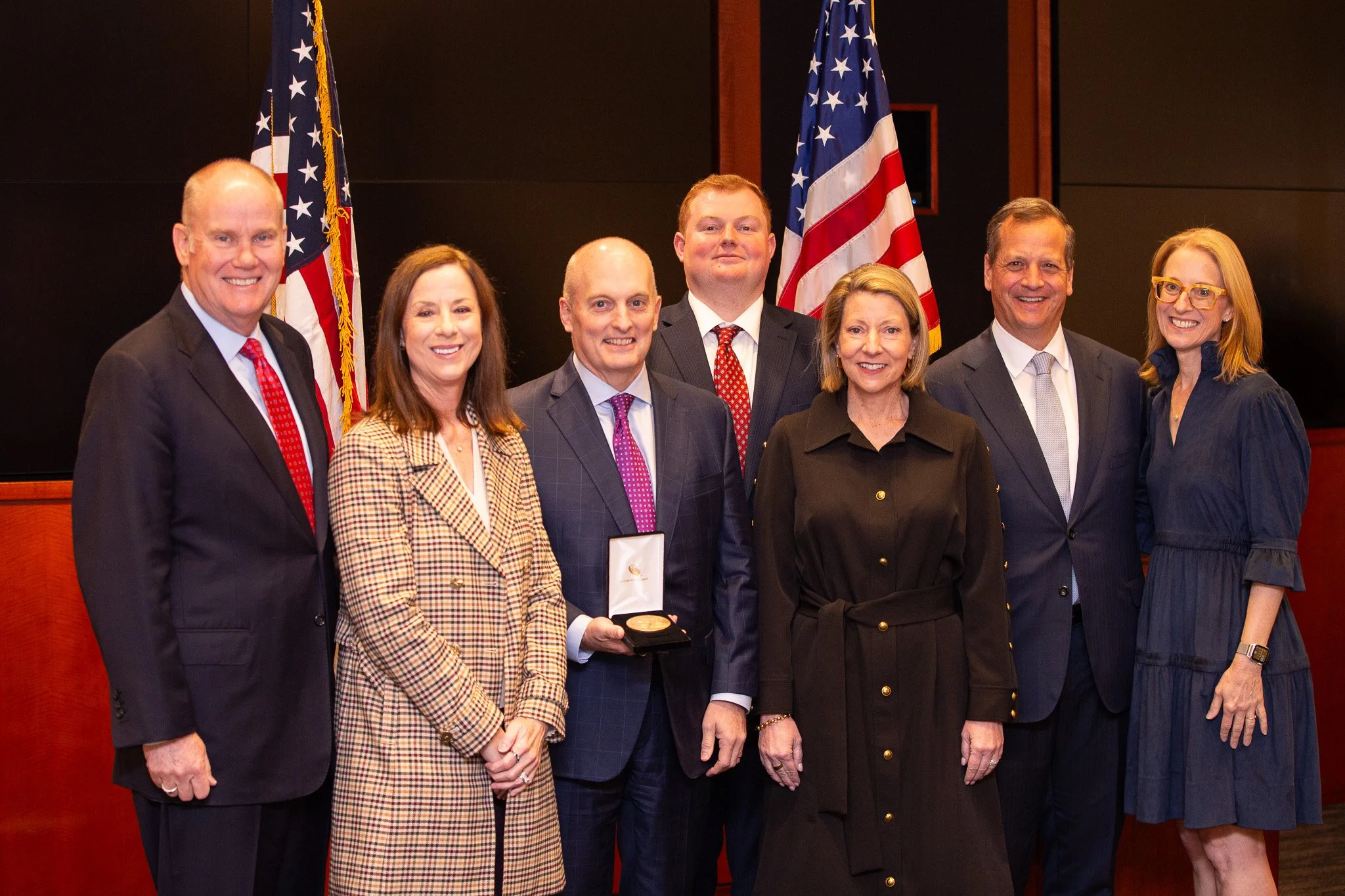 A group of professionals and leaders pose for a formal photograph during a recognition event, standing together in front of American flags and a ceremonial backdrop. The image captures a moment of achievement and collaboration, highlighting the profe