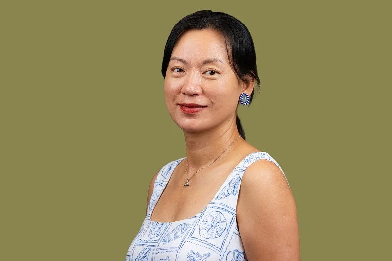 A professional headshot featuring a woman wearing a patterned white and blue top against a muted olive-green background. The portrait showcases a relaxed expression. 