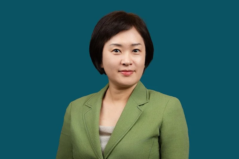 A professional headshot featuring a woman wearing a light green blazer against a deep teal background. The portrait showcases a clean, centered composition. 