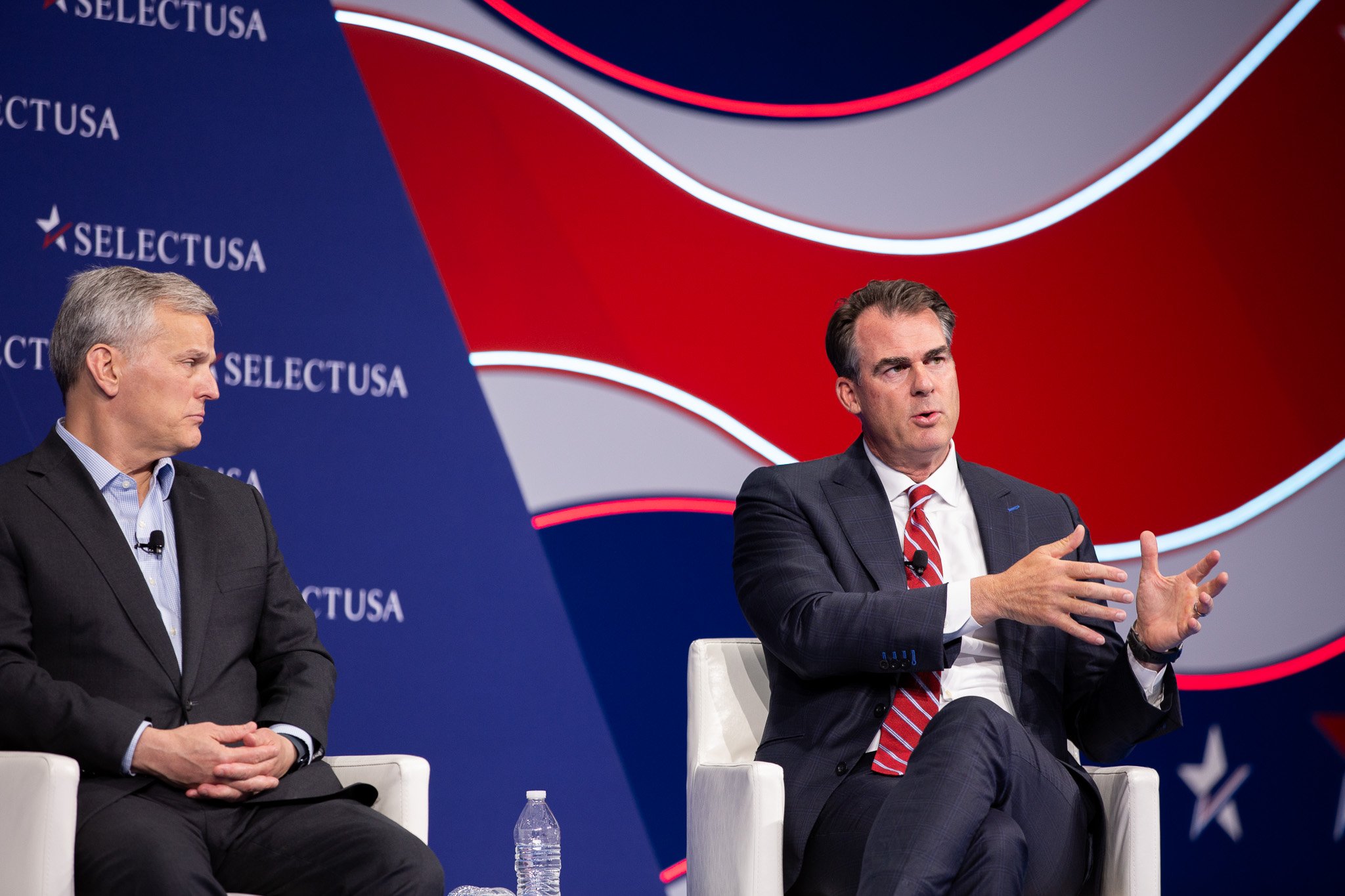 Two executives participate in a panel discussion at the SelectUSA Investment Summit, sharing insights on business investment and economic growth. Set against the event's distinctive red, white, and blue stage design, the discussion captures a dynamic